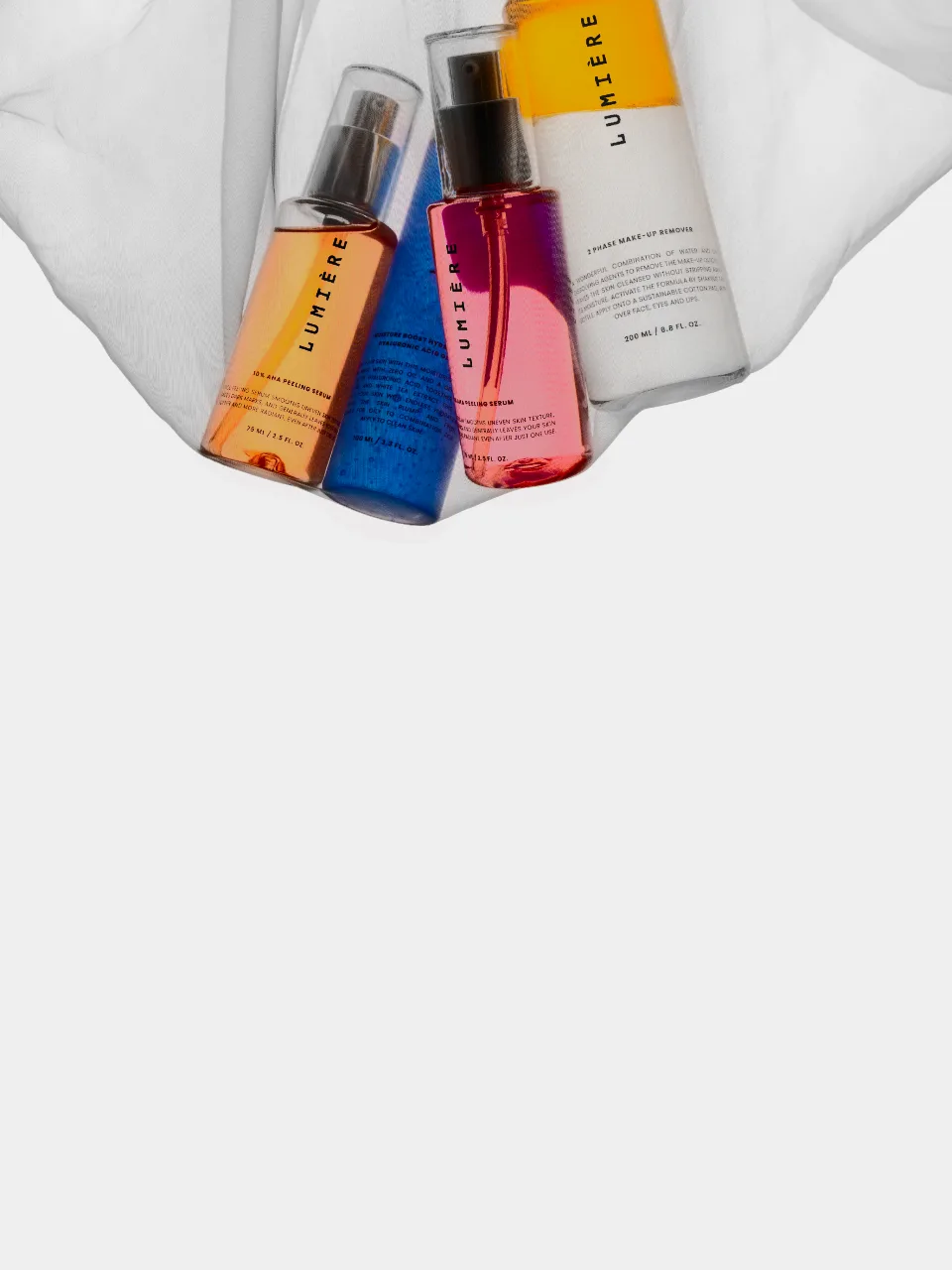 Three skincare bottles (a spray, a blue-capped dropper bottle, and a pink-orange gradient bottle) nestled in draped white fabric