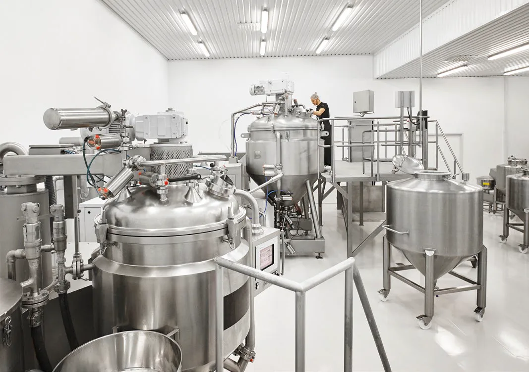 Interior of a cosmetics manufacturing facility with large stainless-steel mixing tanks and a technician working in the background