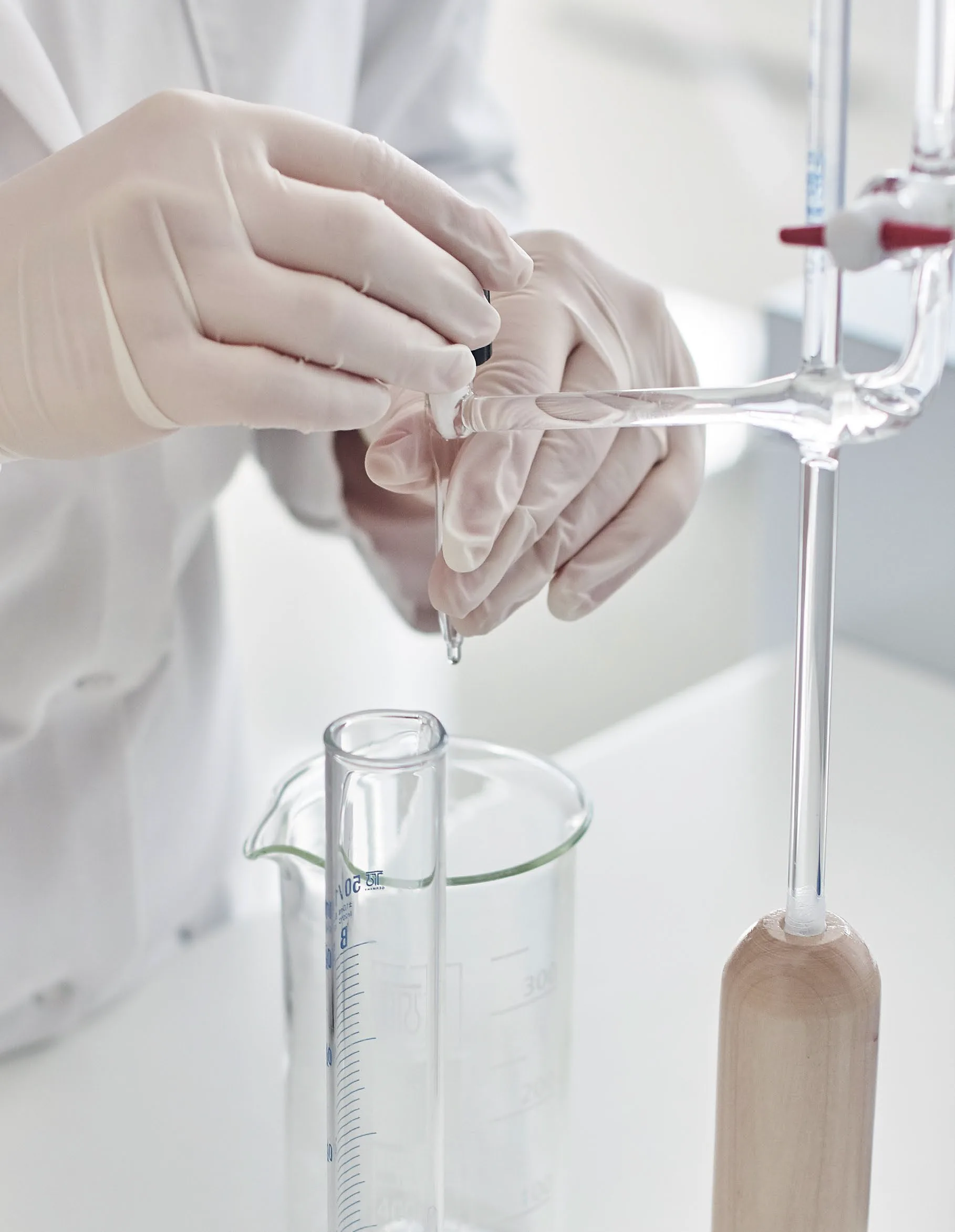 Gloved lab technician's hands holding a pipette over a glass beaker, with a glass stirring rod in the background