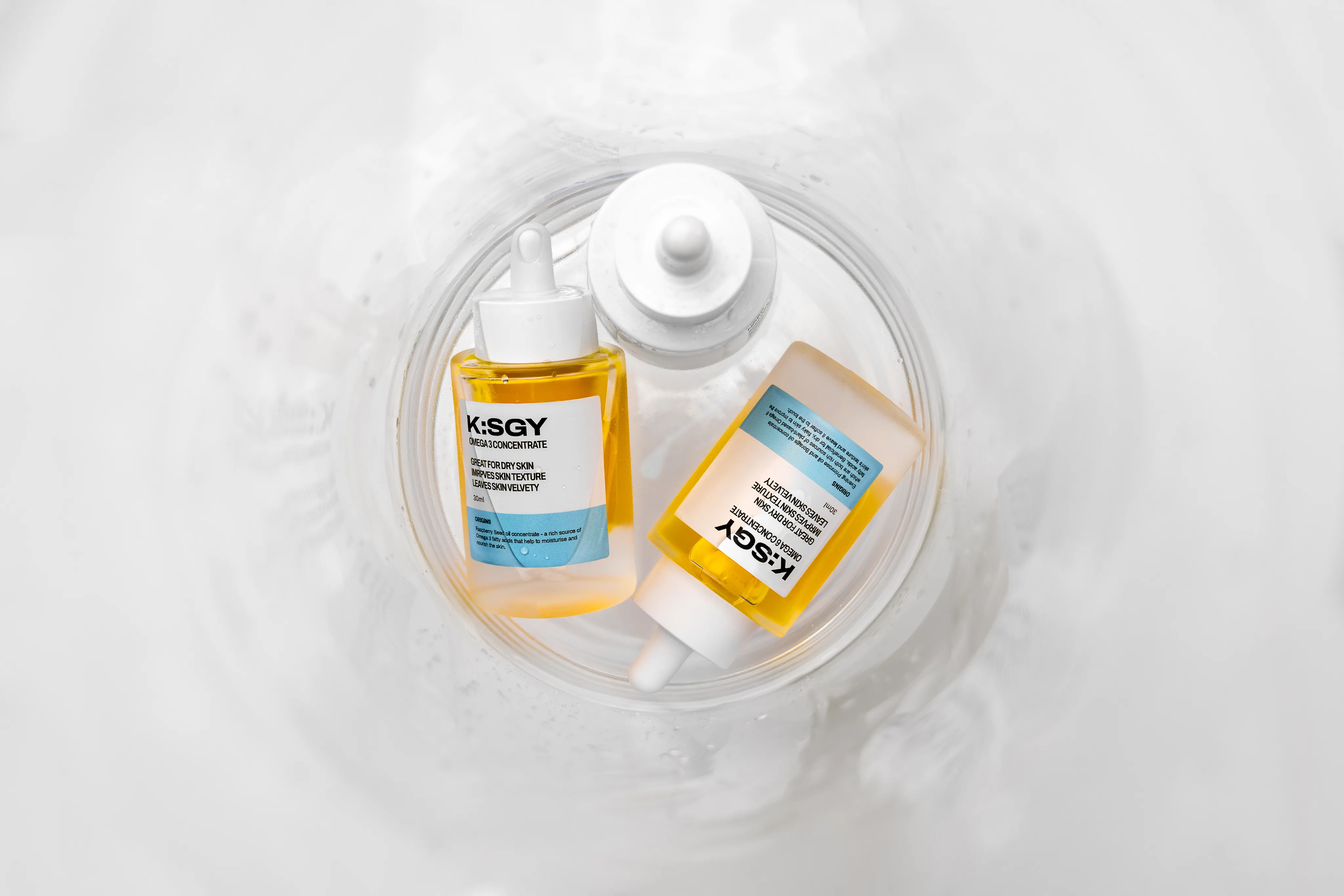Two K:SGY skincare dropper bottles arranged in a soft white swirl of fabric